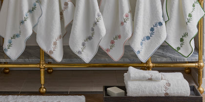 White Daphne towels with floral vine embroidery in assorted colors, hanging on a brass rack.