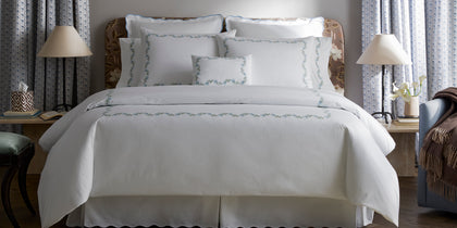 White Daphne bedding with blue floral embroidered border, styled on a bed near a floral headboard.