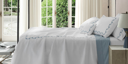 White Daphne Matelasse quilted bedding with blue floral embroidery, styled on a bed near French doors.
