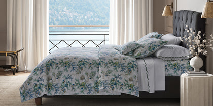 Del Tesoro coral and shell print bedding in blue and green, styled on a bed overlooking a lake.