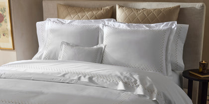 White Dorothy bedding with polka-dot embroidered border, styled on a bed with gold quilted pillows.