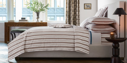 Brown and white striped Elio bedding styled on a bed near a patterned curtain.