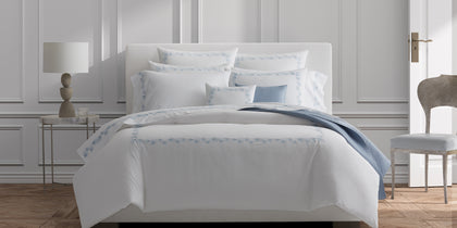 White Feather bedding with blue embroidered trim, styled on a bed with a blue throw.
