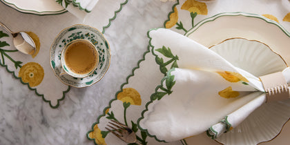 Gold Gisele placemat and napkin with a floral border, set on a marble table with china.