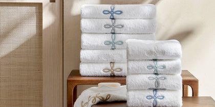 White Gordian Knot towels stacked in a bathroom with embroidered borders in blue and gold.