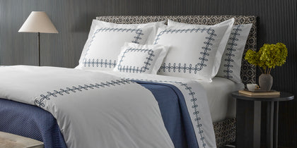 White Gordian Knot bedding with a navy embroidered border and navy throw, styled on a bed.