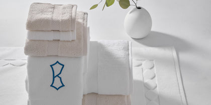 Stack of white and tan Guesthouse towels with a navy monogram, beside a bath mat and a bud vase.