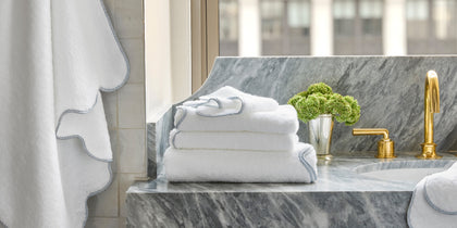 White Jasper towels with blue braided trim on a gray marble vanity with a gold faucet.