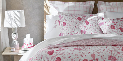 Pink Khilana floral print bedding with a plaid sham, styled on a bed with a caned headboard.