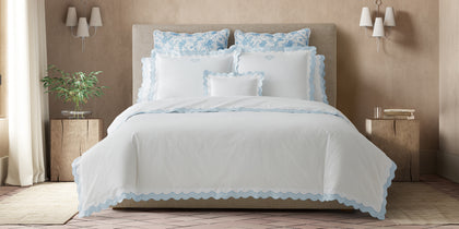 White Lorelei scalloped bedding with light blue trim, styled on a bed with floral shams.