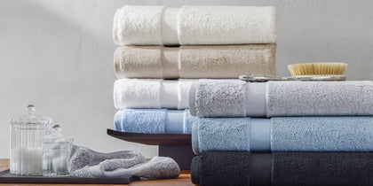 Lotus towels stacked in ivory, tan, blue, and black, with a bath brush on a wooden dresser.