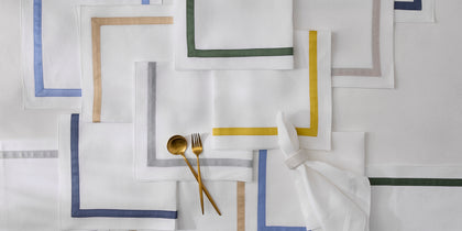 White Lowell napkins with bordered trim in assorted colors, arranged flat with gold flatware.
