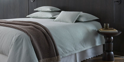 Sage-striped Matteo bedding with a brown waffle throw, styled on a bed against a dark paneled headboard.