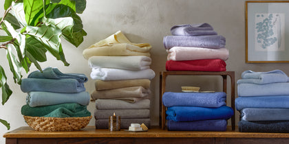 Milagro bath towels stacked in multiple colors on a wood console beside a leafy plant.
