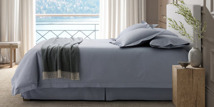 Gray Milano Hemstitch bedding with a gray throw, styled on a bed beside a lakeside window.