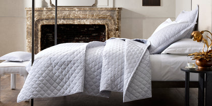 White diamond-quilted Milano Quilt bedding styled on a bed beside a marble fireplace.