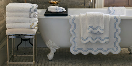 White Mirasol towels with scalloped blue trim draped on a clawfoot tub.