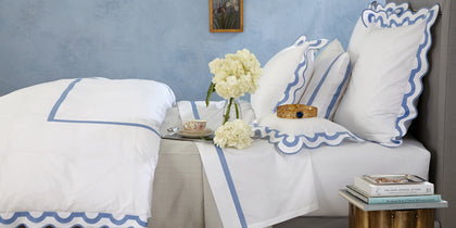 White Mirasol bedding with scalloped blue trim on a blue upholstered bed with white hydrangeas.