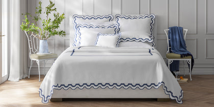 White Mirasol Matelasse coverlet with navy scalloped trim on a bed between two white iron chairs.
