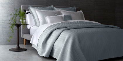Gray herringbone-quilted Netto bedding styled on an upholstered bed beside a round side table.