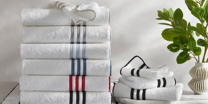 White Newport towels with striped borders in assorted colors stacked beside folded towels and a plant.