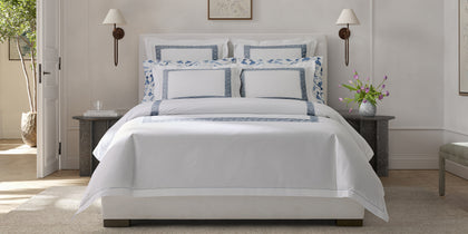 White Ophelia bedding with blue Greek key trim on a bed between stone side tables.