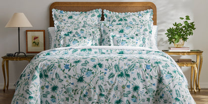 White Palmyra floral-print bedding on a bed with a cane headboard and gold side tables.