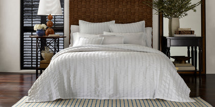White seersucker-textured Panama bedding on a bed with a woven headboard and striped rug.