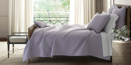 Lavender diamond-quilted Petra bedding styled on a bed near a window with a cane bench.