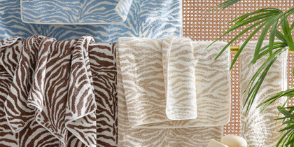Zebra-print Regine towels in blue, brown, and tan draped beside a caned wall and palm leaves.