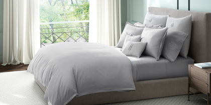 White Talita Satin Stitch bedding with monogrammed pillows on a bed beside a balcony window.
