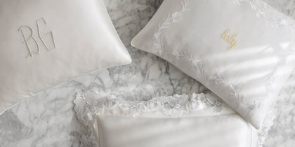 White silk pillows from The Silk Collection with a monogram, floral embroidery, and lace trim on marble.
