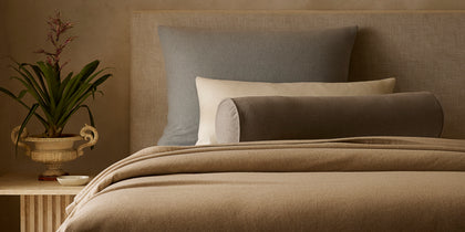 Tan Venus wool blanket and pillows styled on a bed beside a potted plant.