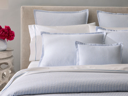 Blue-striped Matteo bedding styled on an upholstered bed with red peonies on a stone side table.