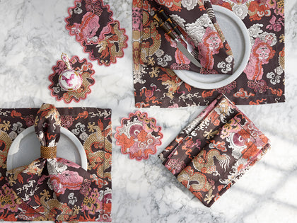 Brown Magic Mountain dragon print placemats, napkins, and scalloped coasters, arranged flat on marble.