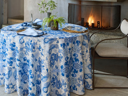 Blue and white Magic Mountain dragon print tablecloth set near a fireplace, with woven placemats and a folded napkin.